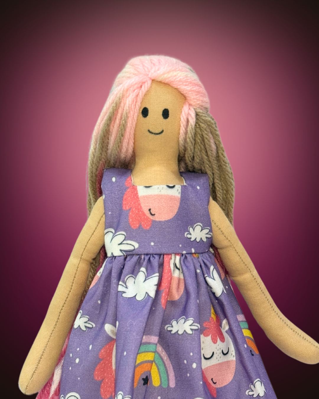Light-skinned soft rag doll with pink yarn hair, dressed in a purple unicorn print dress, designed for imaginative and screen-free play.