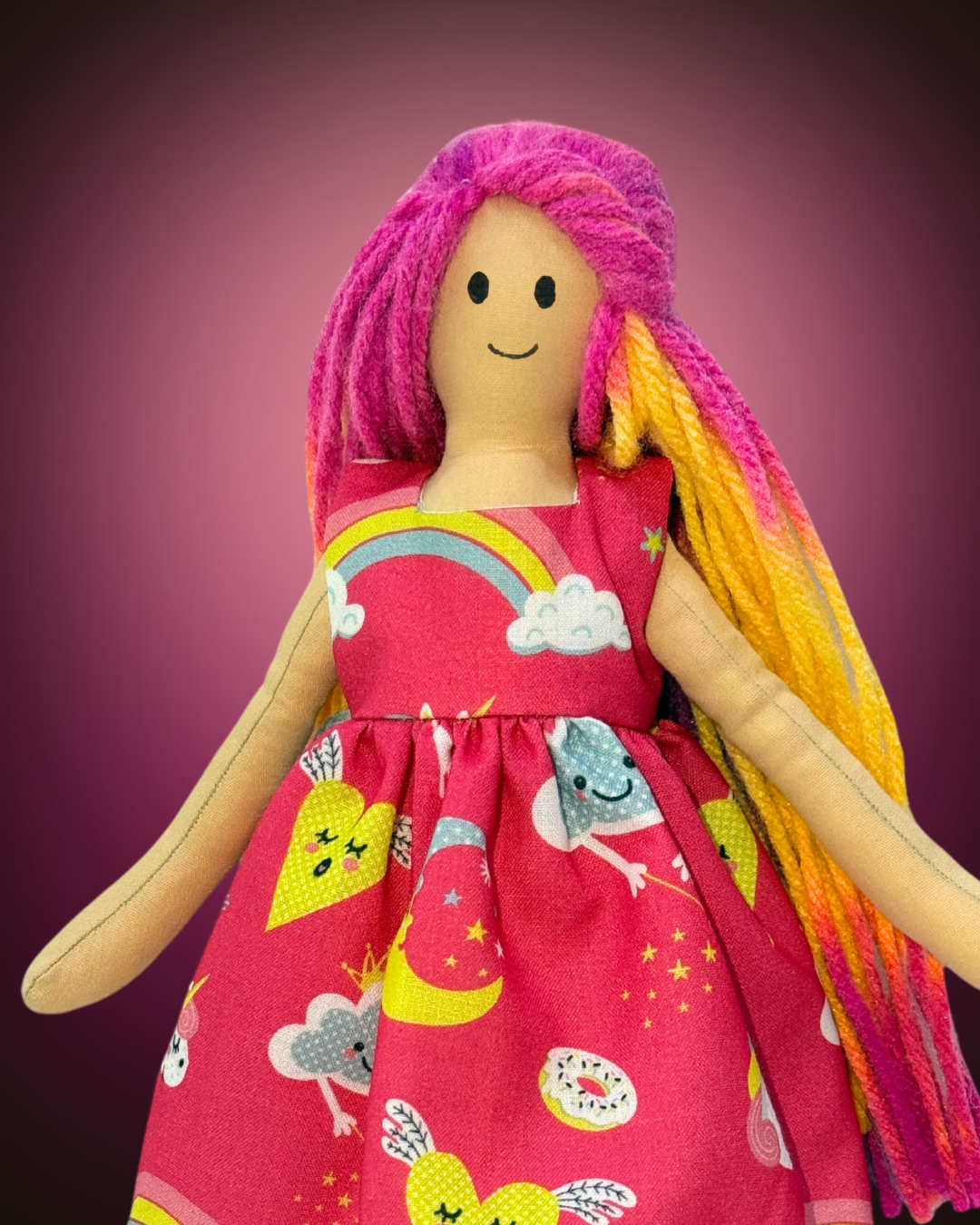 Soft handmade rag doll with colorful yarn hair, wearing a bright pink dress with rainbows and clouds, ideal for sustainable gifting